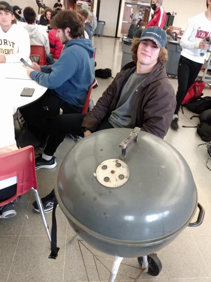 A boy sits next to a grill he brought for the occasion.  