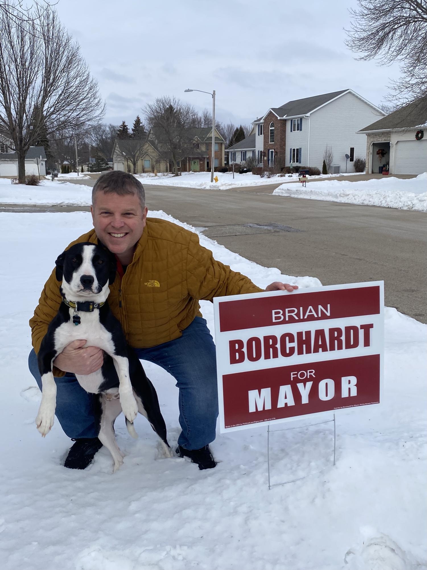 Brian Borchardt crowching, posing next to a sign that reads "Brian Borchardt for Mayor" with his dog. 